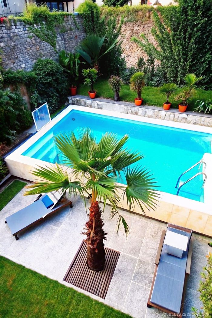 Elegant above ground pool with blue water surrounded by stone walls, tropical palm tree, lounge chair, and lush greenery creating a serene outdoor oasis.