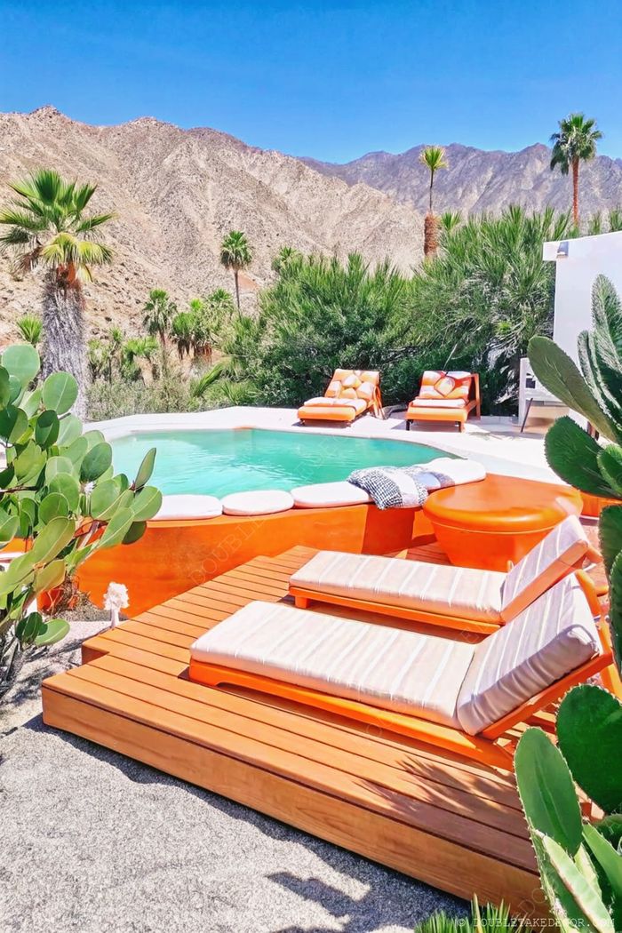 Above ground pool with orange rim surrounded by tropical plants, mountain views, and stylish orange lounge chairs on wooden deck.