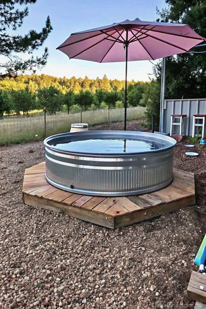 Elevated galvanized tub pool on wooden deck with pink umbrella, surrounded by trees and rustic outdoor entertaining space.