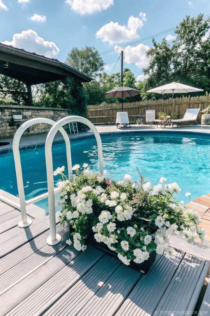 A serene above ground pool with a white ladder, surrounded by a wooden deck, flowers, sun loungers and umbrellas for relaxation.