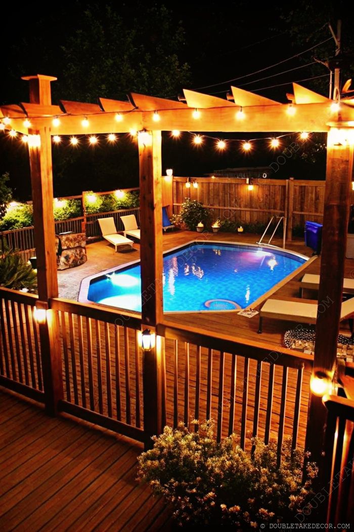 Illuminated above ground pool with wooden deck and pergola adorned with string lights, creating a cozy nighttime retreat.