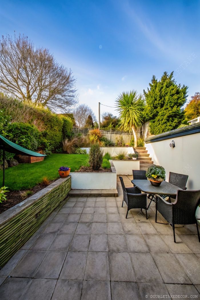 Landscaped garden featuring a gray-tiled patio with wicker dining set, flowing into a tiered garden with ornamental plants and trees.