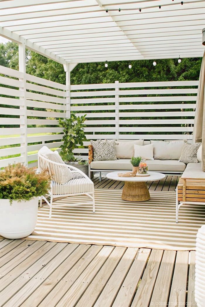 Private outdoor living area with white sectional sofa, wicker chair, and circular coffee table on a wooden deck with potted plants.