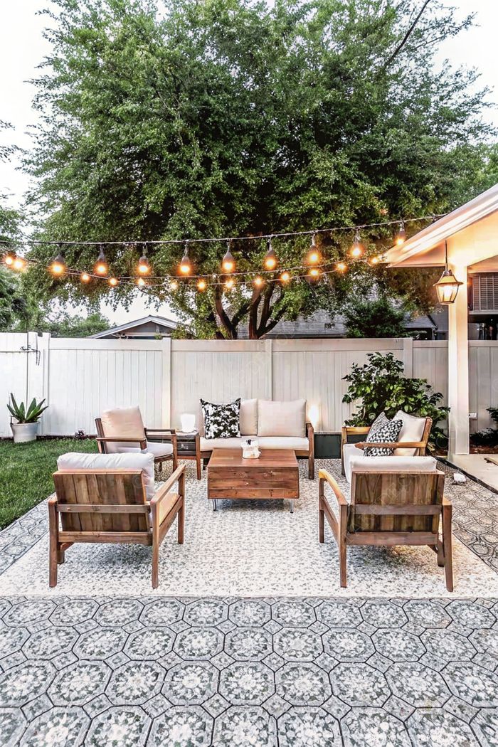 Comfortable wooden chairs with cushions arranged around a coffee table on a patterned rug, under string lights with a backdrop of greenery and white fence.