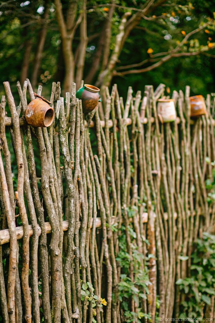Rustic wooden branch fence with decorative clay pots integrated into its structure, surrounded by greenery.