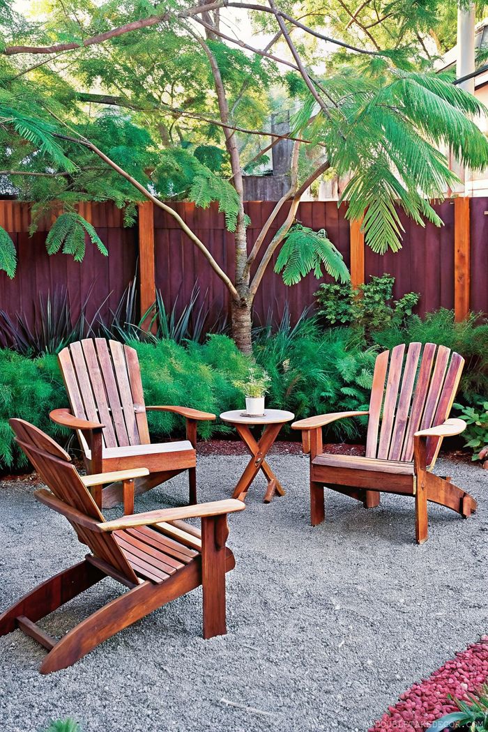 Wooden Adirondack chairs around a table on gravel, surrounded by lush foliage and a burgundy fence for privacy.