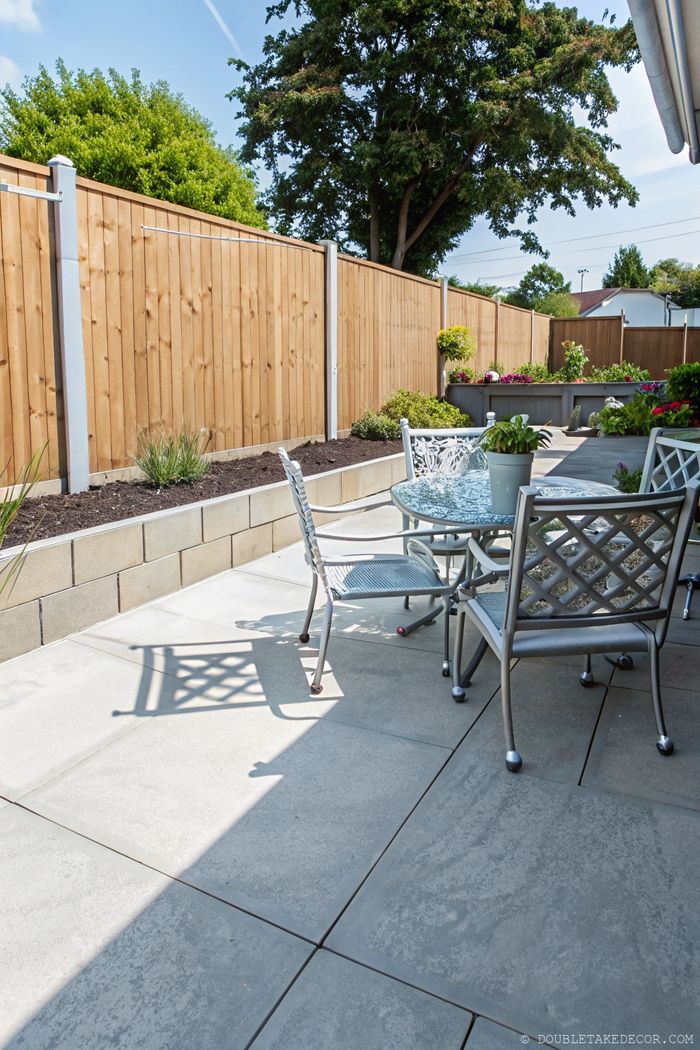 Secluded patio with round glass table, decorative metal chairs, and privacy fence surrounded by colorful garden beds
