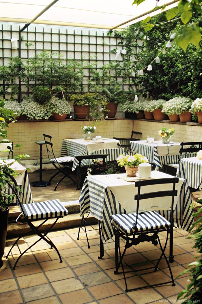 Outdoor dining area with lattice privacy wall adorned with greenery, featuring navy-striped tablecloths and ambient string lights