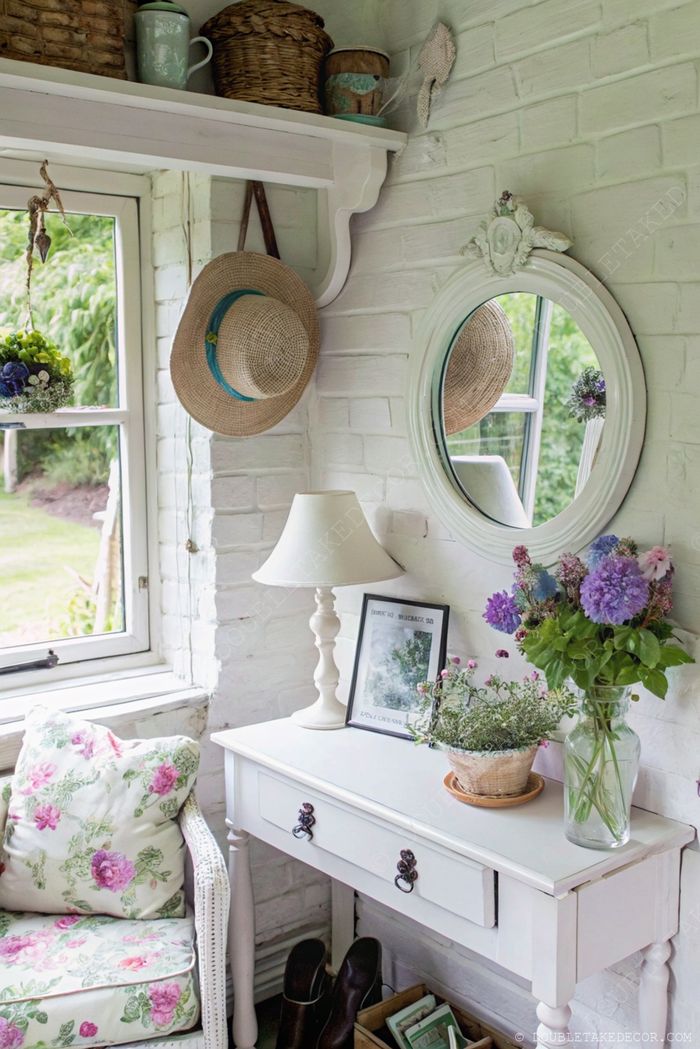 Niche de brique blanche charmante avec console, miroir, fleurs violettes et fauteuil à coussins fleuris pour une ambiance cottage sereine.