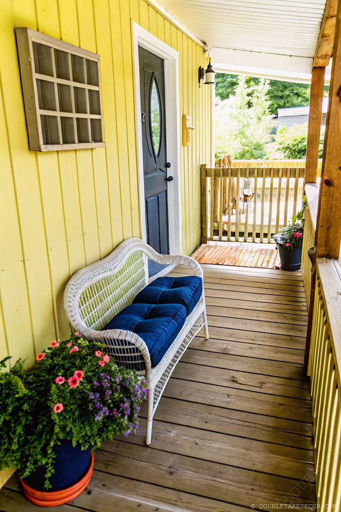 Chaleureux porche jaune de cottage avec banc en osier blanc, coussins bleus et jardinières colorées sur plancher bois