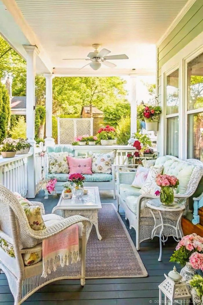 Porche de cottage cosy avec meubles en osier blanc, coussins colorés, pots de fleurs et ventilateur de plafond pour une vraie détente au jardin.
