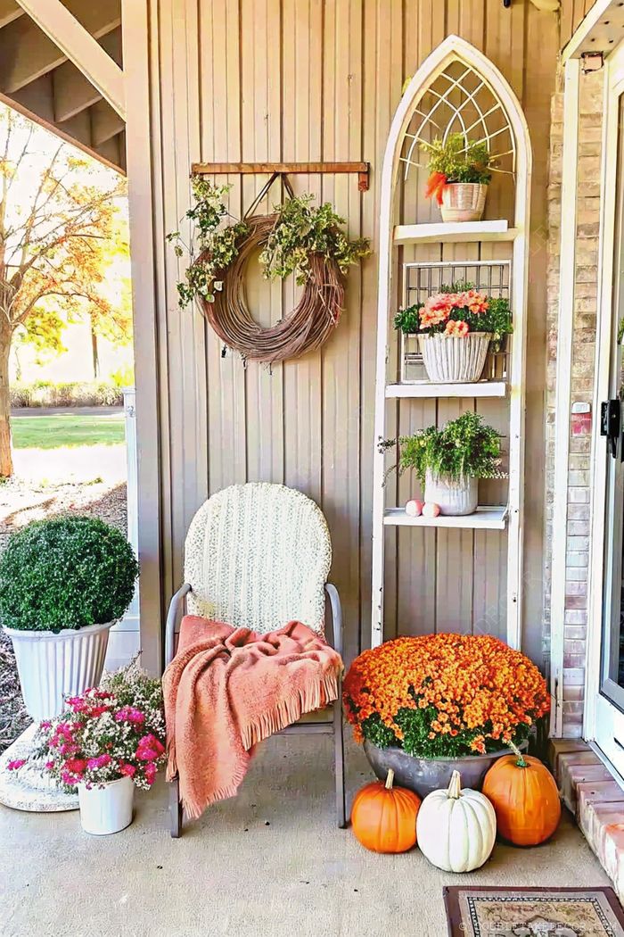 Porche accueillant avec fauteuil en osier, plaid orange, chrysanthèmes, couronne rustique, et citrouilles créant une ambiance chaleureuse.