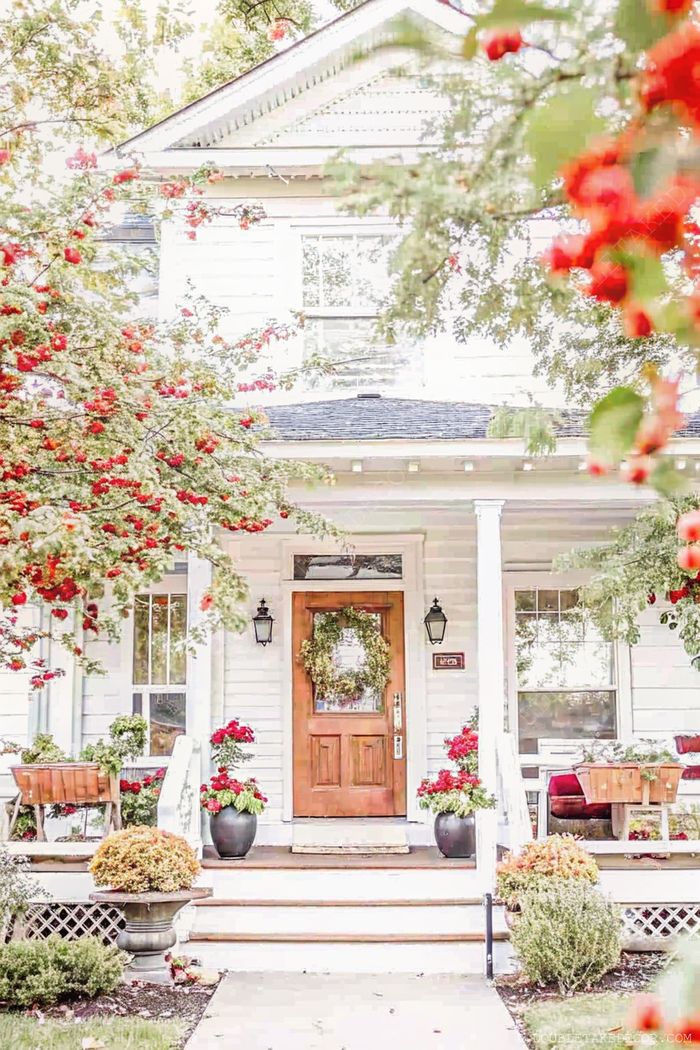 Cottage blanc accueillant avec porte en bois, couronne de saison, appliques symétriques, fleurs rouges et banc créant une ambiance chaleureuse.