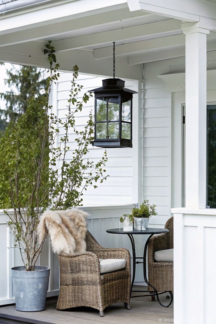 Porche de cottage charmant avec deux fauteuils tressés, coussins crème, plaid imitation fourrure, et petite table noire sous une lanterne suspendue.