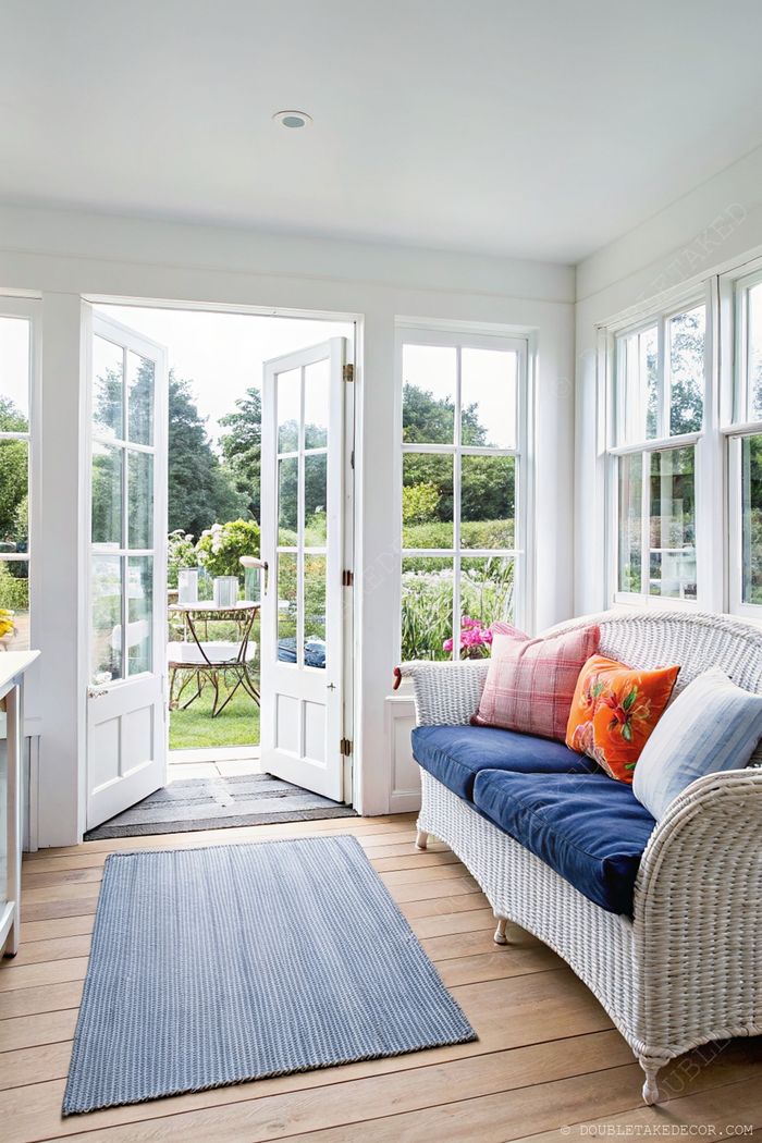 Véranda blanche lumineuse avec canapé en osier, coussins colorés et portes ouvertes sur un jardin et un coin repas extérieur.