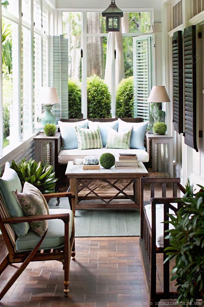 Véranda confortable avec volets blancs, canapé clair et coussins bleu-vert, fauteuil en bois, table basse rustique et plantes en pots.