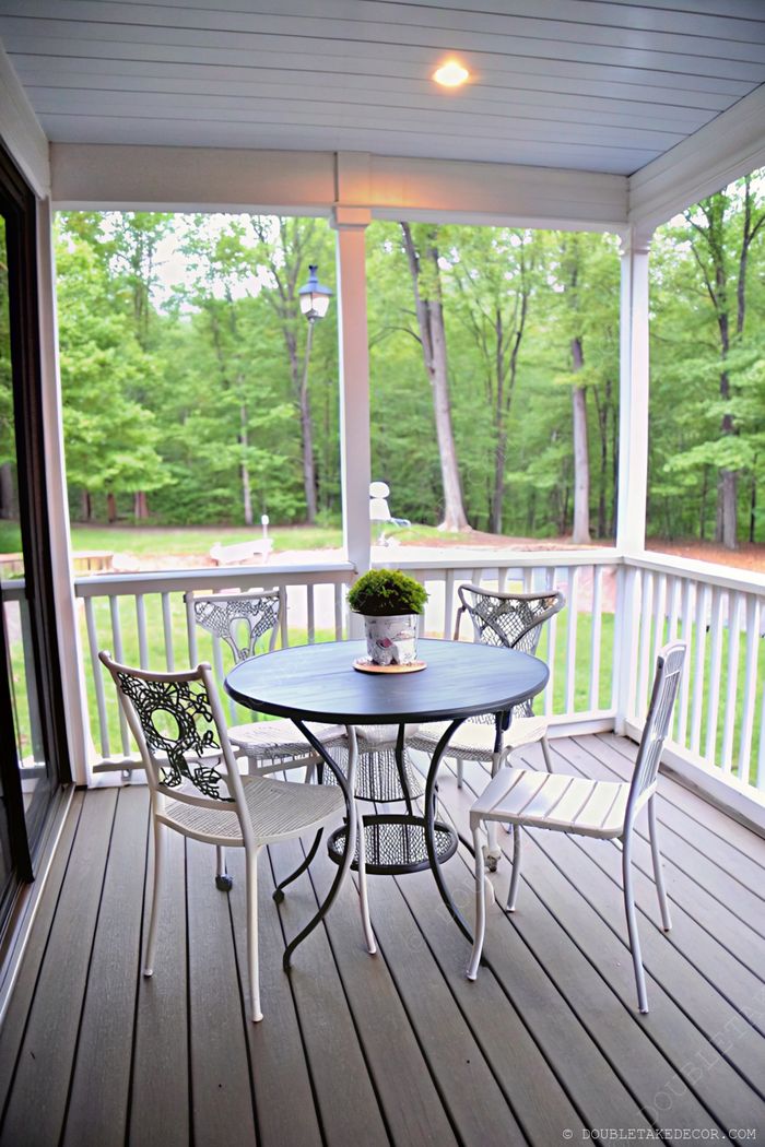 Porche charmant avec grande table ronde noire et quatre chaises blanches en métal, terrasse en bois et vue sur le jardin protégée par une balustrade.