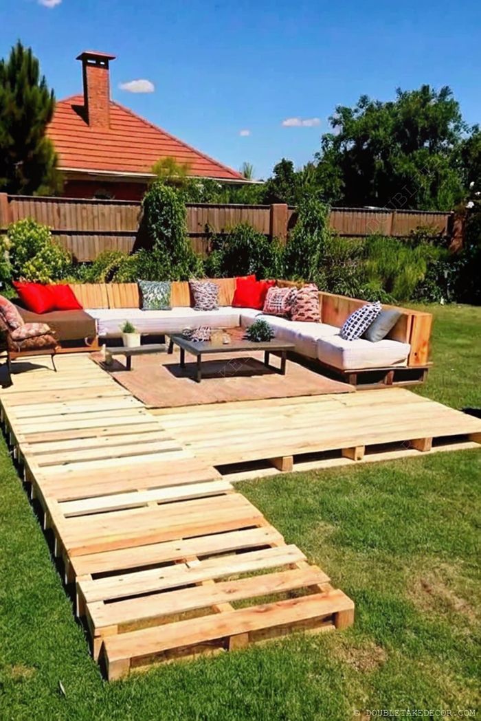 Terrasse DIY en palettes avec canapé d’angle, coussins clairs, coussins rouges et table basse assortie dans un jardin.