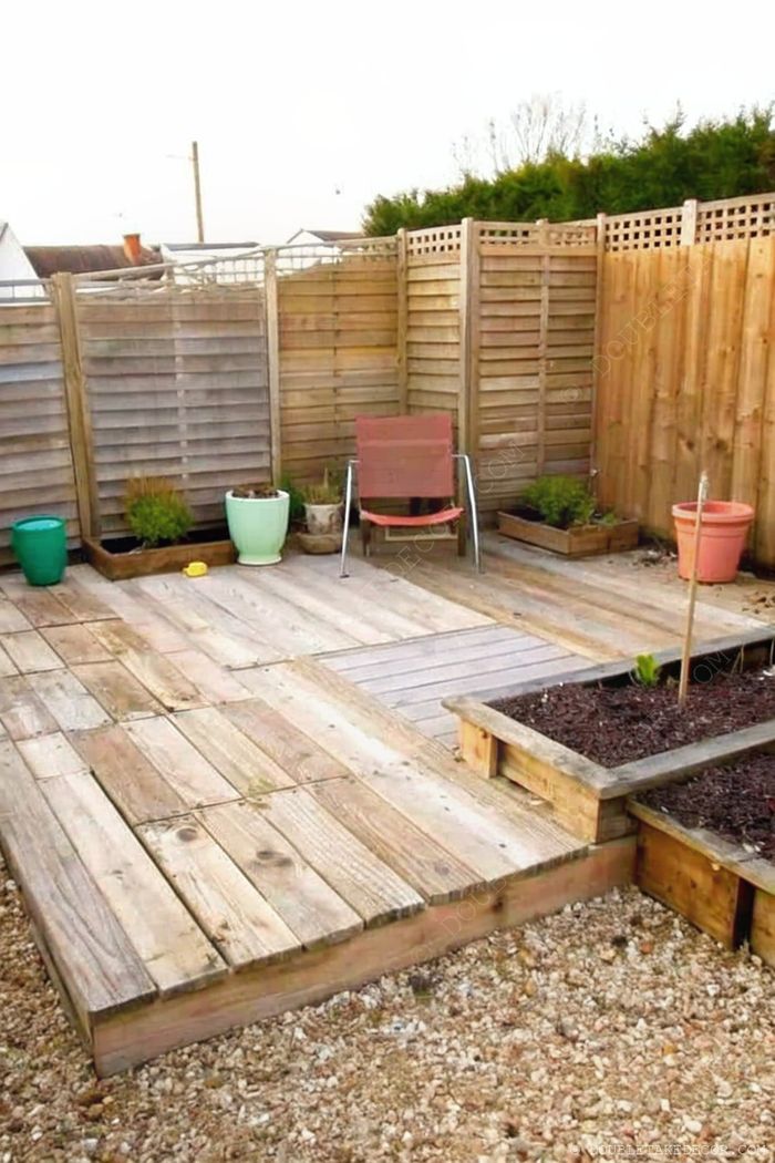 Petit patio extérieur en bois, clôture pour l’intimité, chaise pliante, et jardinières colorées prêtes pour le jardinage.
