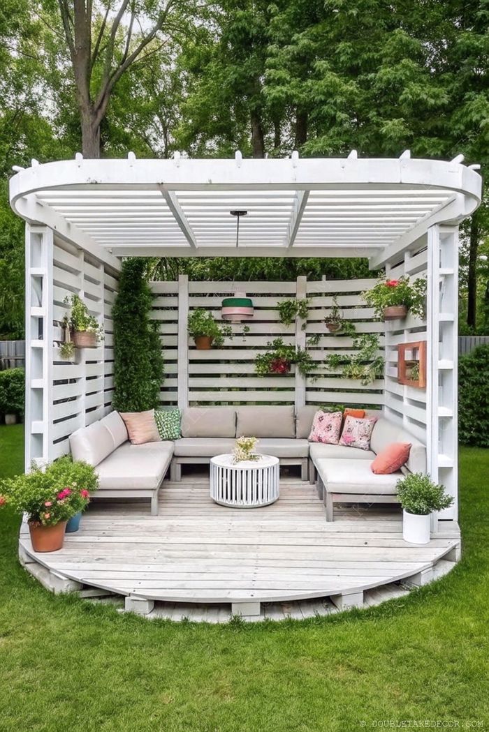 Gazebo moderne en bois blanc avec assise grise en U, table ronde et panneaux de verdure, posé sur terrasse en bois.