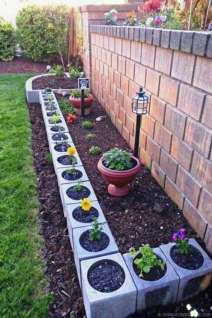 Wiederverwendete Betonsteine bilden eine strukturierte Gartenbegrenzung. Sie sind mit bunten Sonnenblumen und violetten Blüten bepflanzt, dazu passen Tontöpfe und eine dekorative Lampe.