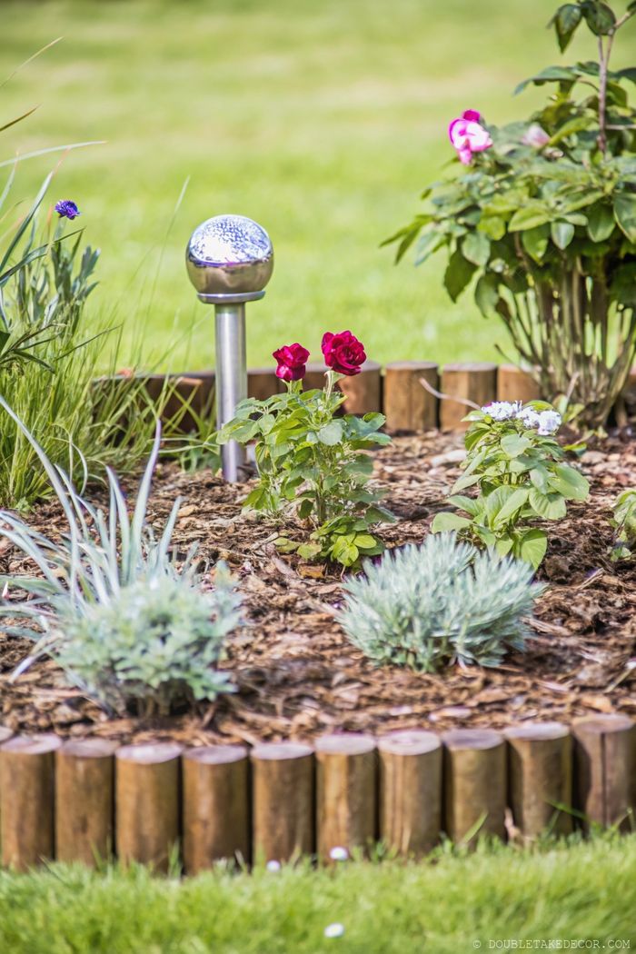 Blumenbeet mit intensiven roten Rosen, umgeben von einer rustikalen Holzeinfassung, dekorativer Gartenbeleuchtung und sattem Grün.