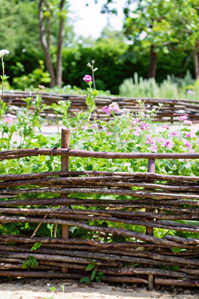 Verflochtener Astzaun, der einen natürlichen Rand um einen lebhaften Garten mit rosa und weißen Blumen bildet, die hindurchwachsen.