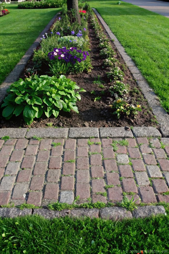 Rechteckiges Blumenbeet, eingefasst mit Steinen und violetten sowie weißen Blüten, dazu ein sauberer Pflasterweg für einen eleganten Gartenrand.