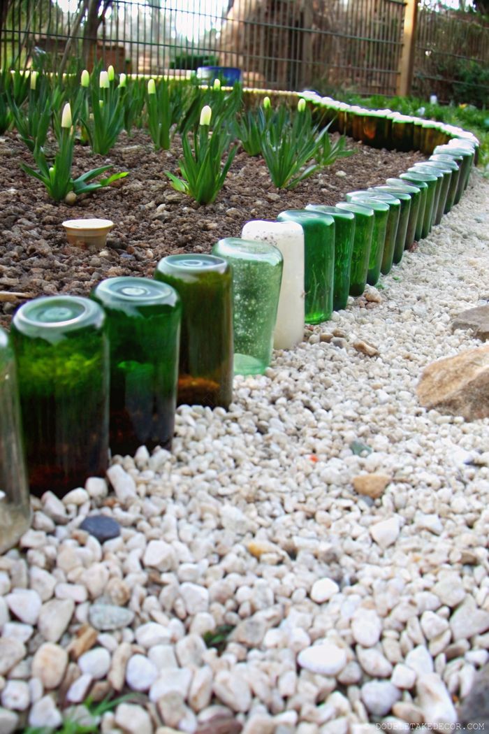Dekorativer Gartenrand aus aufrecht eingesetzten grünen und klaren Glasflaschen, die eine verspielte Kante um kleine Pflanzen auf weißem Kies bilden.