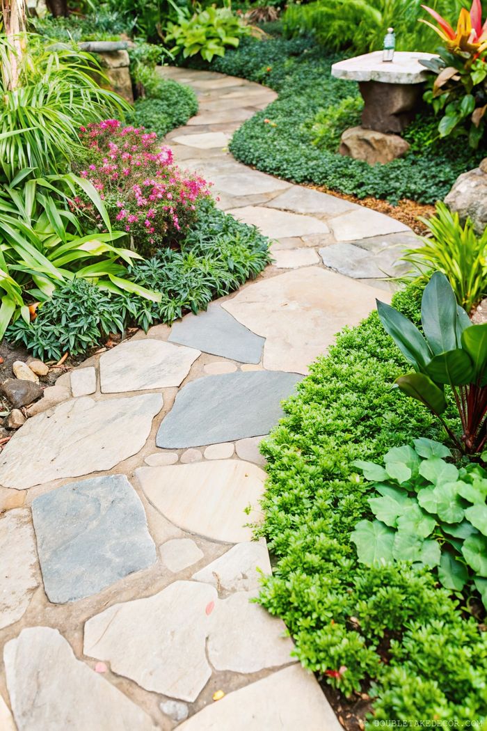 Schöner Gartenweg aus unregelmäßigen Steinplatten, flankiert von vielen Grünpflanzen und rosa Blüten, der zu einer gemütlichen Sitzecke führt.