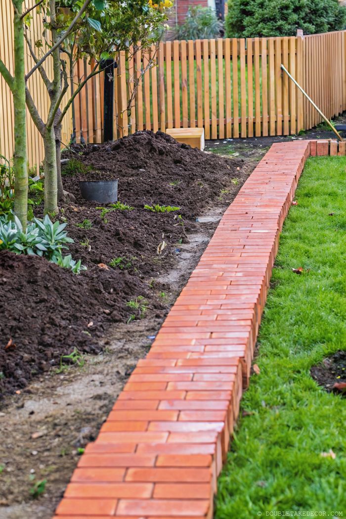 Klar abgegrenzter Gartenweg aus roten Ziegelsteinen, daneben frische Erde, Zierpflanzen und ein Holzzaun als Hintergrund.