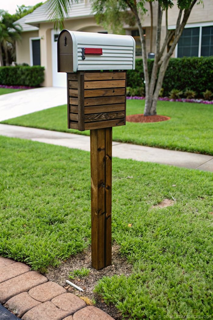 Stylish silver mailbox with red flag mounted on a natural wooden post, surrounded by green lawn and stone pathway.