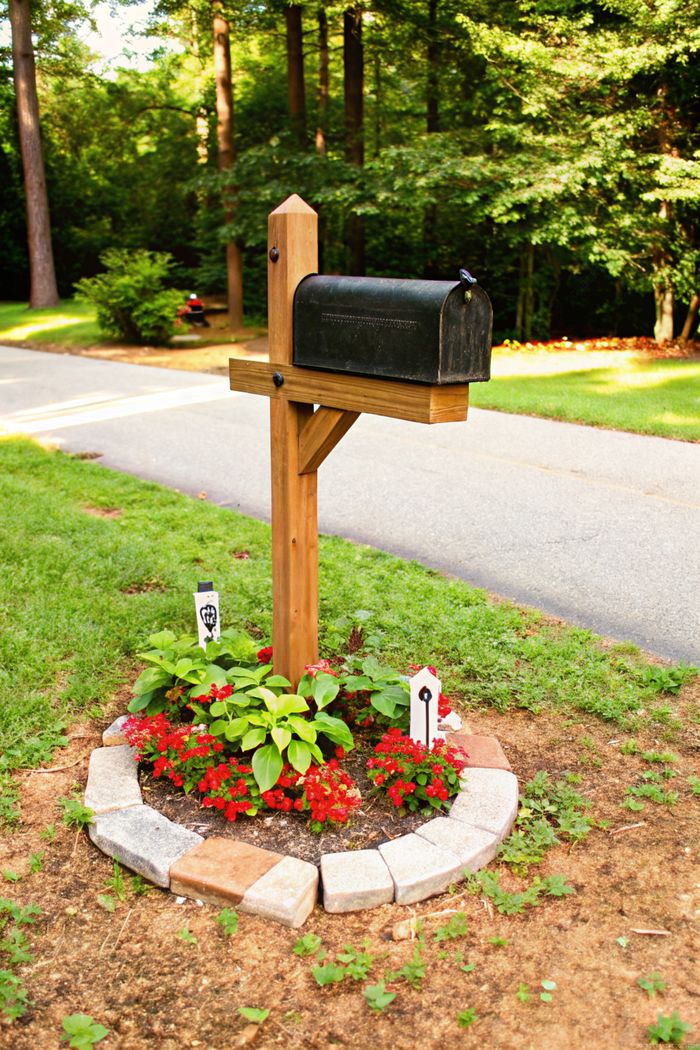 Weathered mailbox on wooden post surrounded by circular flower bed with red blooms and decorative stone border