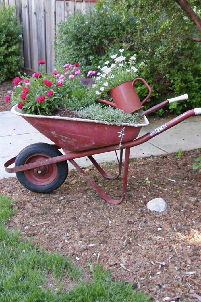 Repurposed red wheelbarrow filled with pink and white flowers, topped with a matching red watering can, creating charming shabby chic garden decor.