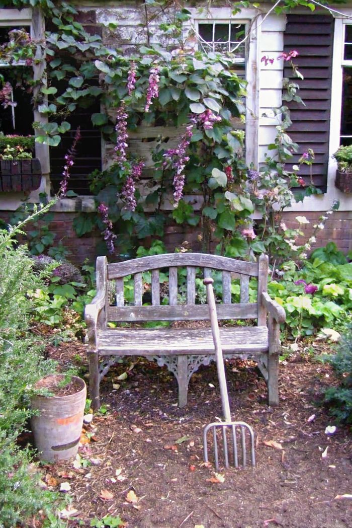 Weathered wooden bench surrounded by lush greenery and blooming flowers, with a gardening fork leaning against it in a charming shabby chic garden setting.
