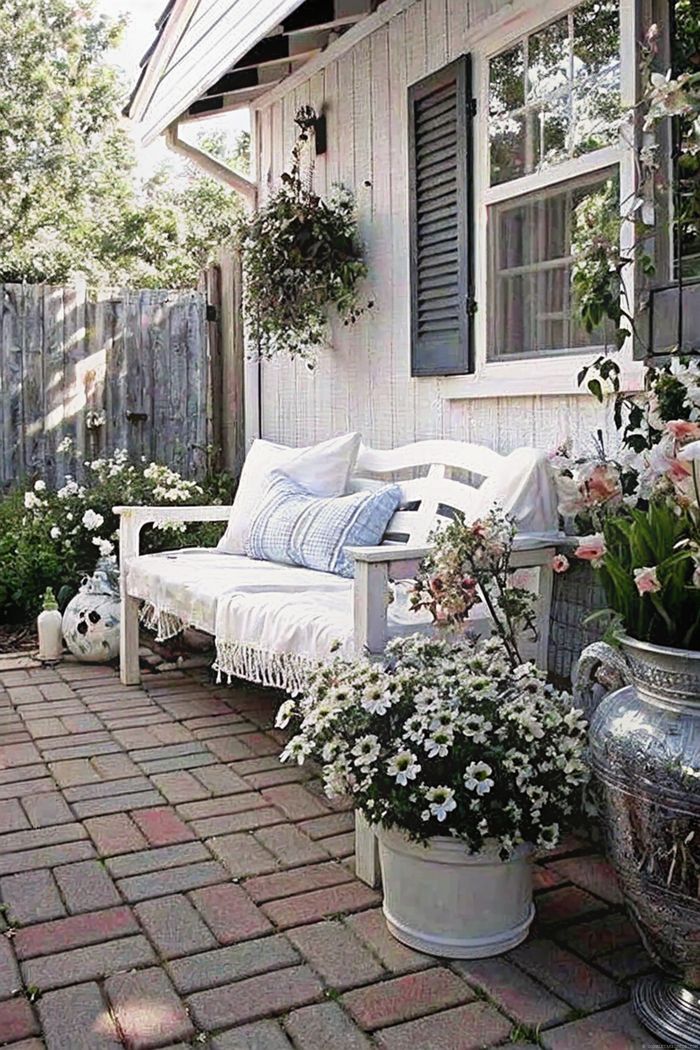 White bench with cushions surrounded by white flowers on a brick patio, featuring decorative vases and rustic wooden fence backdrop.