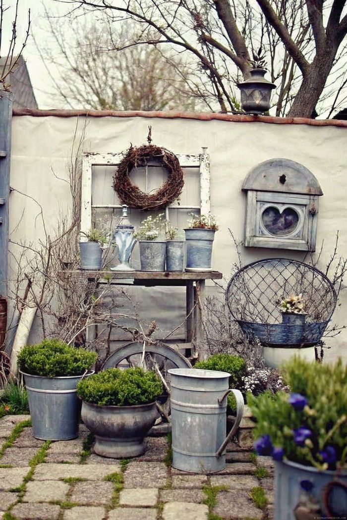 Weathered wall with window and wreath above wooden table displaying galvanized planters, teapot, and herbs on a stone pathway