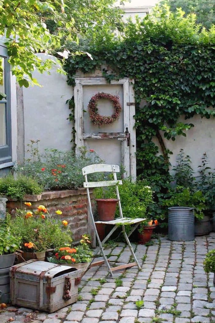 A weathered white chair beside potted blooms, with a vine-covered door and cobblestone pathway creating a serene shabby chic garden retreat.