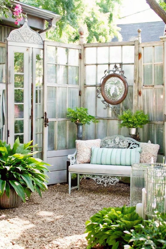 Charming outdoor space with weathered glass doors, vintage metal bench with pastel cushions, surrounded by ferns and a decorative mirror.
