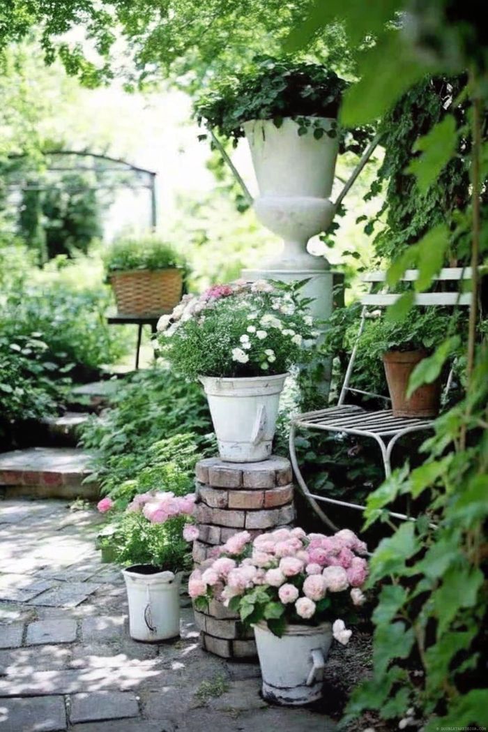 Charming garden with lush greenery, white and pink flowers in rustic containers, and a decorative metal bench along a stone pathway.