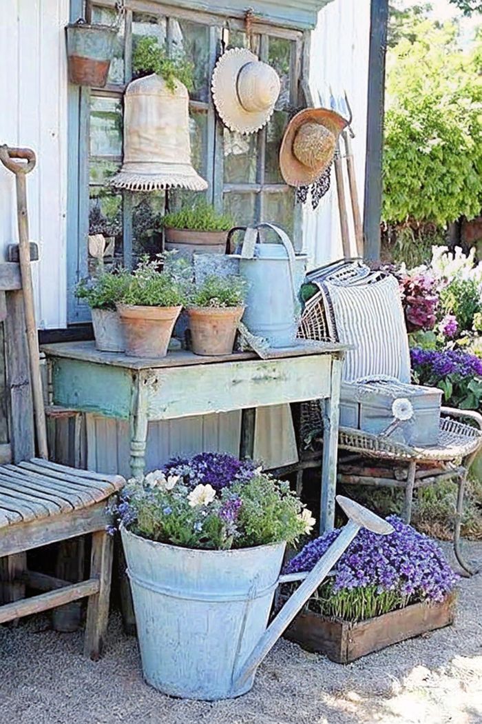 Weathered blue wooden table with terracotta pots, vintage window frame, decorative watering cans, and comfortable seating creating a serene shabby chic garden space.