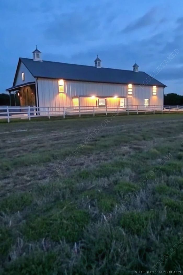 Modern, barndominium stílusú épület fém tetővel, fehér külsővel, meleg fényekkel megvilágítva alkonyatkor, kerítéssel, zöld kerttel körülvéve.