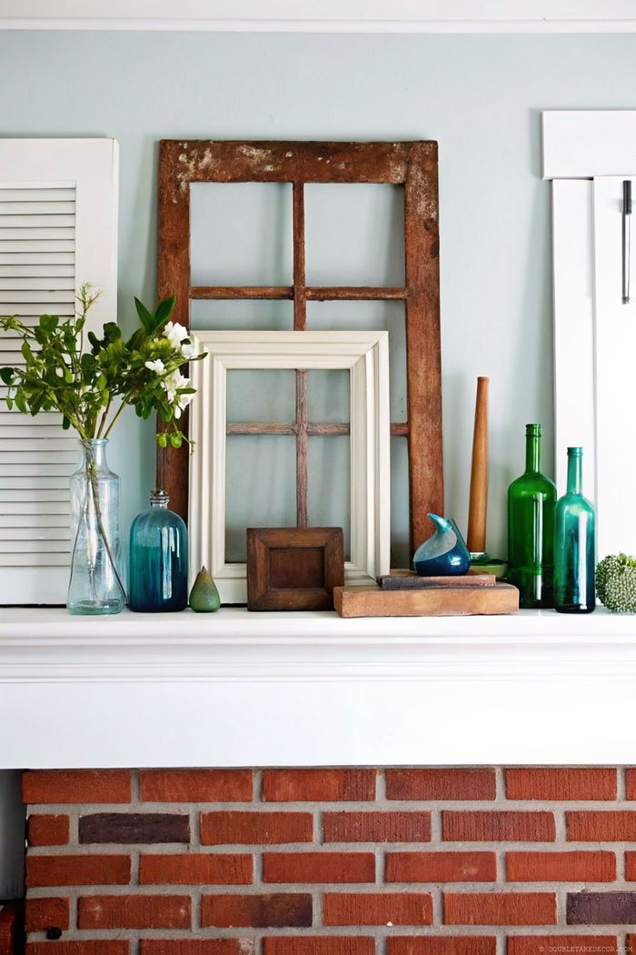 Charmanter Sommerkamin mit Holzfensterrahmen, türkisfarbenen Glasflaschen, frischen Blumen und rustikalen Akzenten auf weißem Sims vor hellblauer Wand.