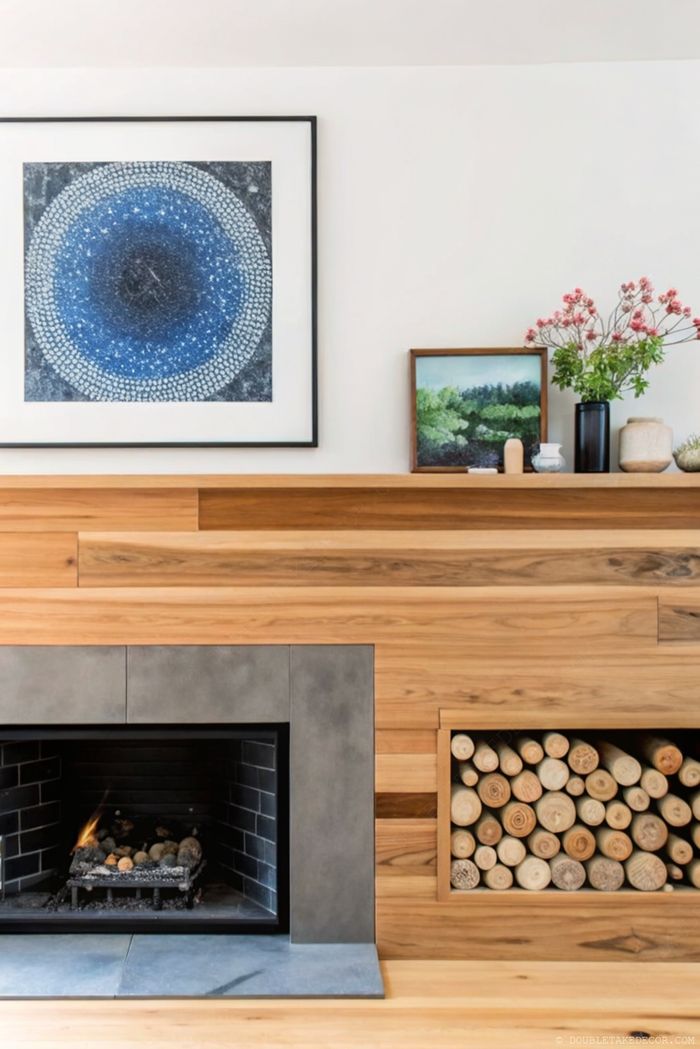 Moderner Kamin mit dunklem Sockel, Kunstwerken in Blau und Landschaft, dahinter Holzwand – klar und natürlich dekoriert.