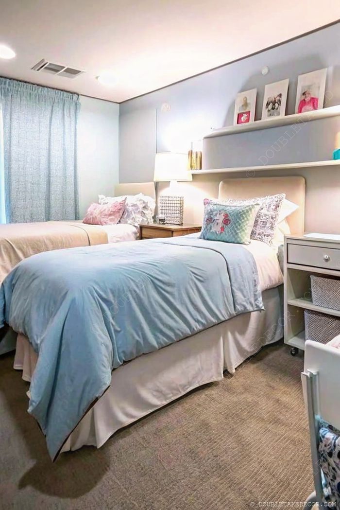 Cozy dorm room with twin beds in blue and beige, natural light, wooden bedside table, and organized storage solutions.