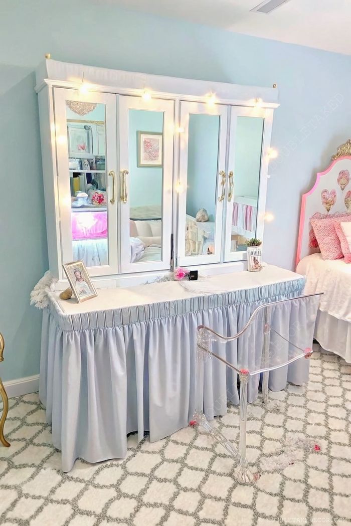 Elegant mirrored vanity with fairy lights, gold handles, blue skirt, and acrylic chair in a soft blue bedroom setting.