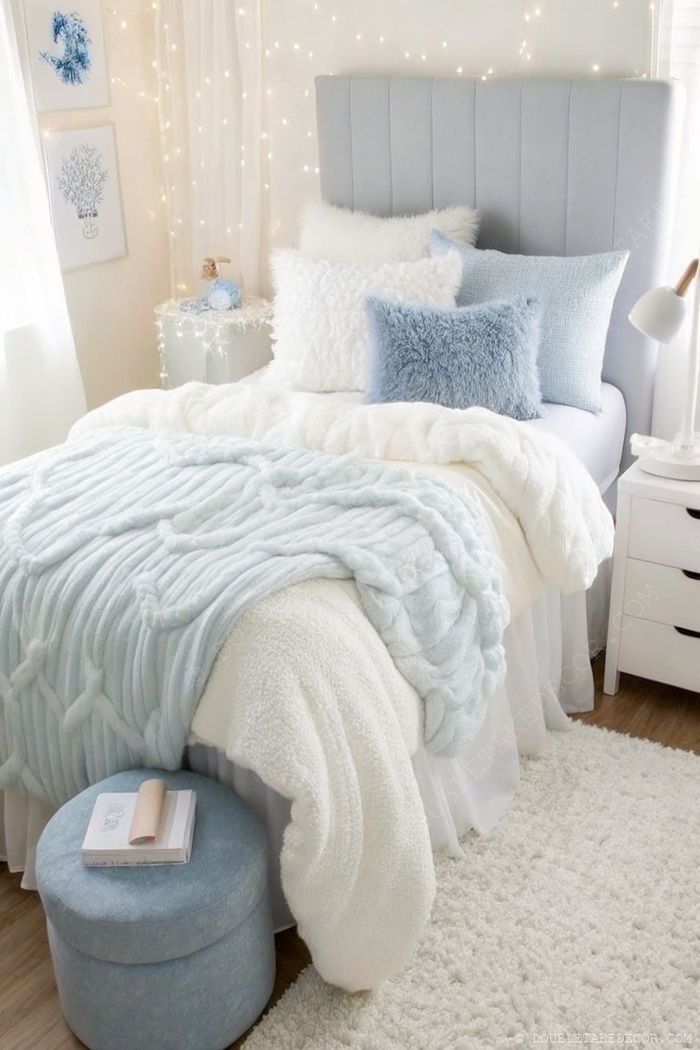 Cozy dorm bedroom with blue-white bedding, textured blankets, small ottoman, fairy lights, and botanical artwork creating a tranquil aesthetic.