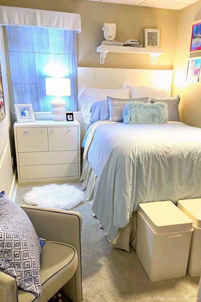 A calming dorm bedroom featuring light beige walls, a blue duvet, minimalist shelf, and cozy reading chair with storage bins.