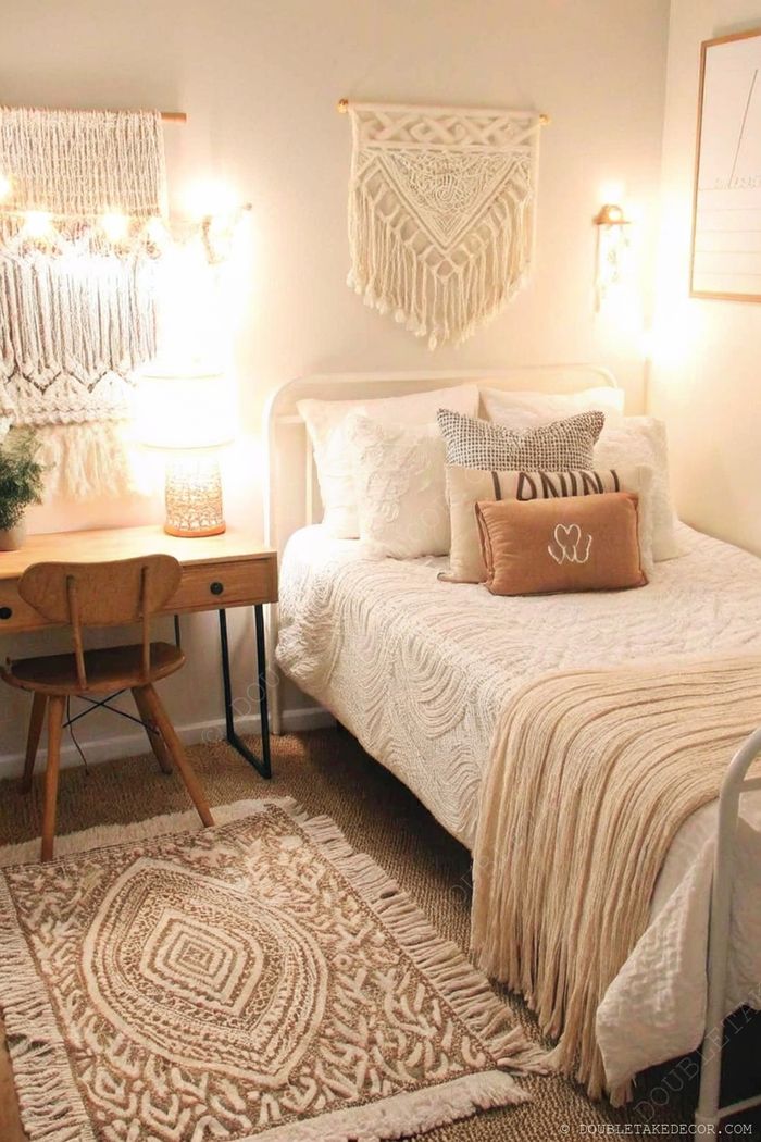 A cozy dorm bedroom featuring a white quilt bed with neutral pillows, wooden desk with woven lamp, and macramé wall hangings with fairy lights.