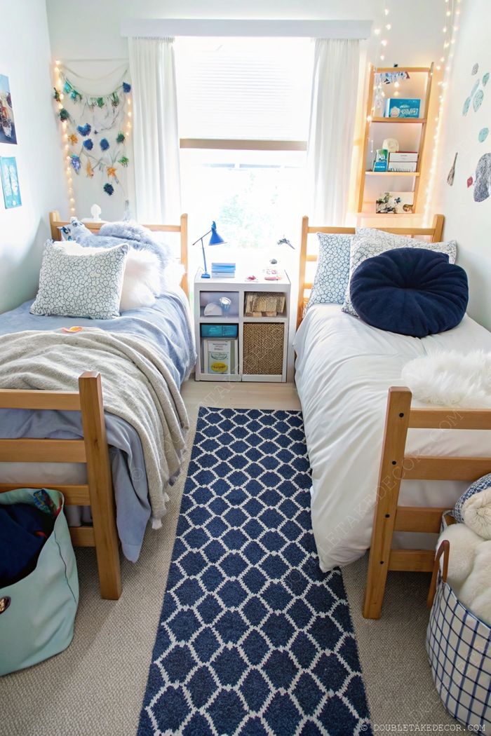 Cozy shared dorm room with twin beds, blue bedding, geometric rug, and central nightstand under soft lighting from window and fairy lights.